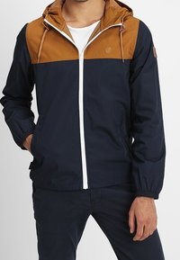 Giacca a vento navy e marrone con zip frontale, cappuccio regolabile e polsini elasticizzati; presenta un logo sul petto e dettagli beige.