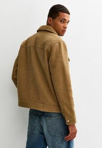 Bruine fleecejas met een kraag, gestructureerd oppervlak en een boxy pasvorm. Voorzien van knoopsluiting aan de manchetten en een rechte zoom, gedragen over denim jeans.