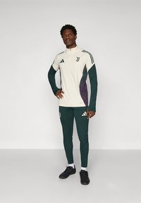 Balta sporta krekls ar zaļiem un violetiem akcentiem, ar ceturtdaļas rāvējslēdzēju un Adidas logotipu; kopā ar pieguļošām zaļām biksēm un melniem apaviem.