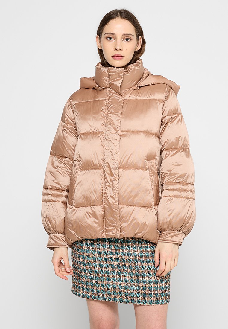 Puchowa, beżowa kurtka z wysokim kołnierzem i kapturem. Ma błyszczącą fakturę i poziome przeszycia. Noszona na wstążkowej spódnicy w kratkę.