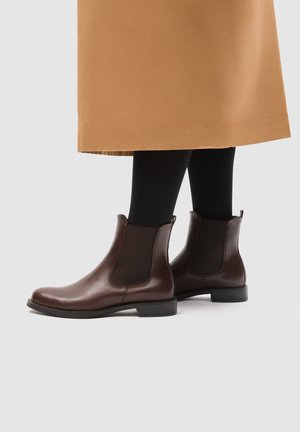 Bottes Chelsea en cuir marron portées avec des collants noirs et une jupe couleur camel, vues de profil sur un fond uni.
