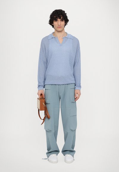 Suéter de punto con textura de color azul claro y cuello, combinado con pantalones cargo de color azul claro. El modelo sostiene un pequeño bolso marrón con correa para el hombro.
