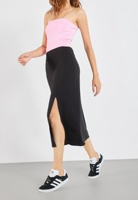 Frau trägt ein pinkfarbenes trägerloses Top, schwarzen Midi-Rock mit seitlichem Schlitz und schwarze Adidas-Sneaker, sie geht vor einem schlichten weißen Hintergrund entlang.