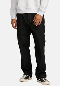 Zwarte spijkerbroek van denim met een relaxed fit, voorzien van contrasterende oranje stiksels, zijzakken en omgeslagen zoom, gedragen met zwart-witte sneakers.
