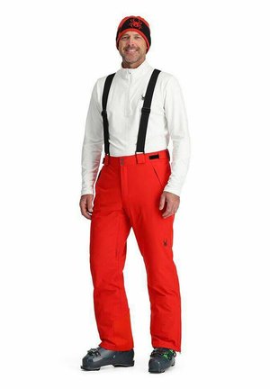 Rote isolierte Skihose mit Hosenträgern, kombiniert mit einem weißen Langarmshirt und einem schwarzen Hut mit einem roten Spinnenlogo.