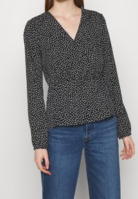 Blouse noire à col en V, manches longues et design péplum. Elle est ornée d'un motif de cœurs blancs sur toute la surface et présente une texture de tissu léger.