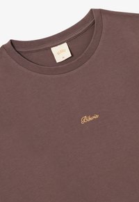 Brun t-skjorte med rund hals, laget av bomull, med et gullbrodert logo "Blkvias" på brystet. Størrelse XL-merke synlig.