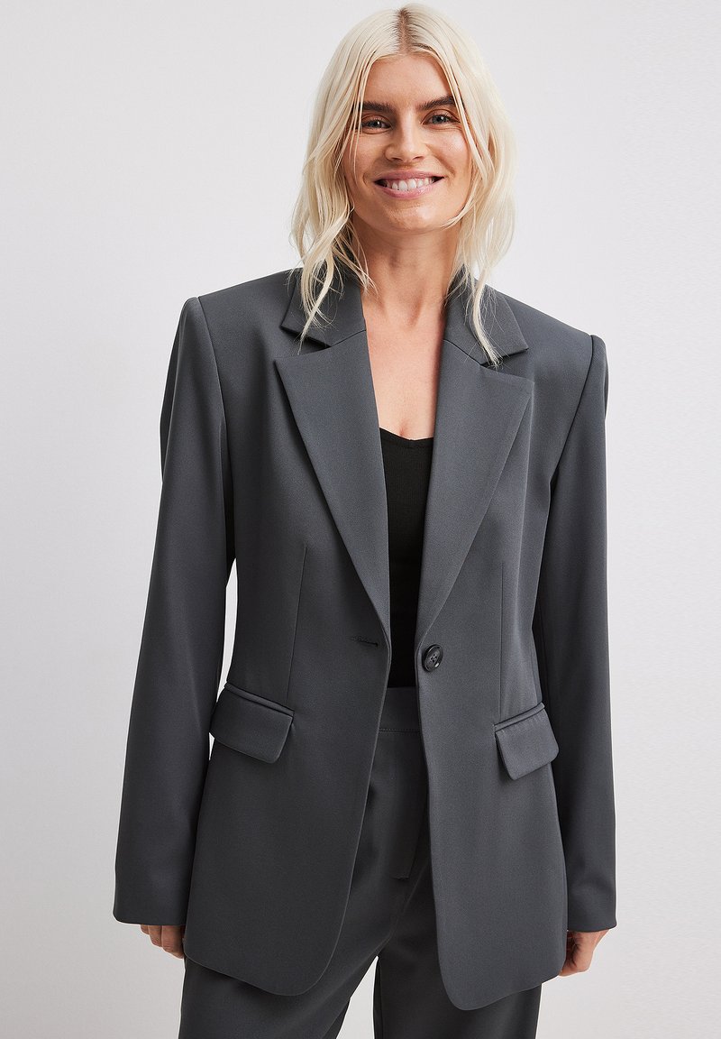 Grå blazer med notch-lapel, enkelt knaplukning, og to frontlommer. Glat tekstur og skræddersyet pasform. Båret over en sort top.