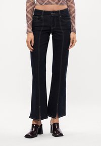 Prilagojeni temno denim kavbojke s sprednjimi šivi, zadrgo na robu in zlato nitko. Kombinirane s sijajnimi maroon petami.