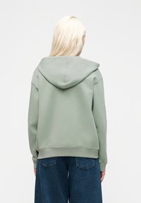 Personne aux cheveux blonds longs portant un sweat à capuche vert sauge et un pantalon en denim bleu foncé, vue de dos sur un fond blanc uni.