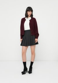 Burgundská vlněná cardigan s knoflíky, nošený přes bílý top, spárovaný s černou skládací sukní a černými lodičkami s mašlemi.
