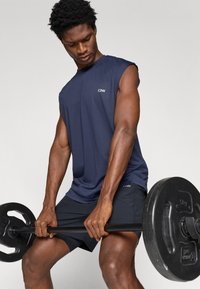 Mornarska brezrokavnik iz športnega materiala z teksturo, v kombinaciji s črnimi kratkimi hlačami. Oseba dviga težo in prikazuje mišičaste roke.