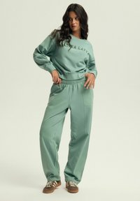 Miętowa bluzka dresowa z nadrukowanym tekstem, w połączeniu z pasującymi spodniami o luźnym kroju. Materiał wydaje się miękki; posiada ściągacze przy mankietach oraz elastyczny pasek w talii.