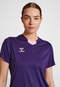 Lilla atletisk skjorte med korte ærmer og v-hals. Har pink detaljer på kraven og skuldrene, samt et pink logo på brystet.