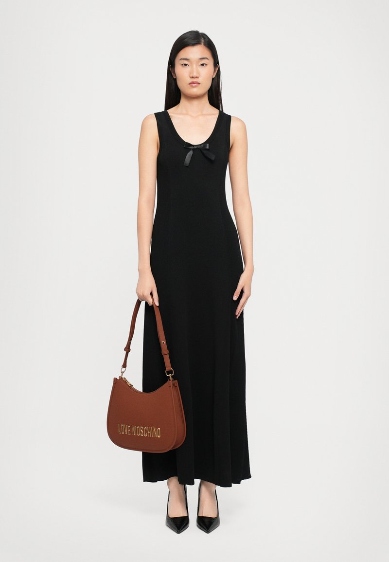 Mulher com um vestido longo preto sem mangas, adornado com um pequeno laço, segurando uma mala castanha "Love Moschino" e usando saltos pretos de bico.