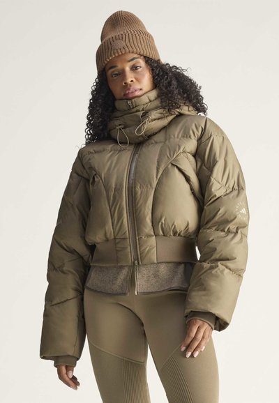Mujer que lleva un gorro de punto marrón, chaqueta acolchada verde oliva con cuello alto y leggings a juego sobre un fondo liso claro.