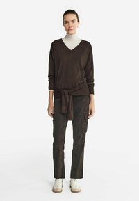 Brauner Strick-V-Ausschnitt-Pullover mit einer gebundenen Taillenpartie, kombiniert mit dunklen Cargo-Hosen und weißen Sneakers vor einem schlichten weißen Hintergrund.