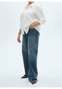 Weißes Leinenhemd mit Knöpfen und einer Tasche, kombiniert mit blauen weiter geschnittenen Jeans und spitzen braunen Schuhen. Das Outfit hat eine lässige Passform.