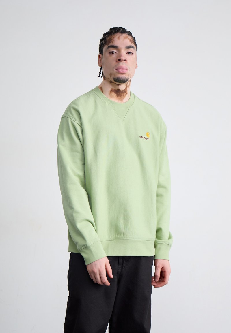 Lys grøn sweatshirt med rund halsudskæring, ribstrikkede manchetter og kant; har et lille broderet logo i gul og rød på brystet.
