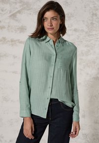 Camicia button-up verde chiaro con righe verticali, una singola tasca sul petto e maniche lunghe arrotolate sui polsi. Realizzata in un tessuto morbido e textured.
