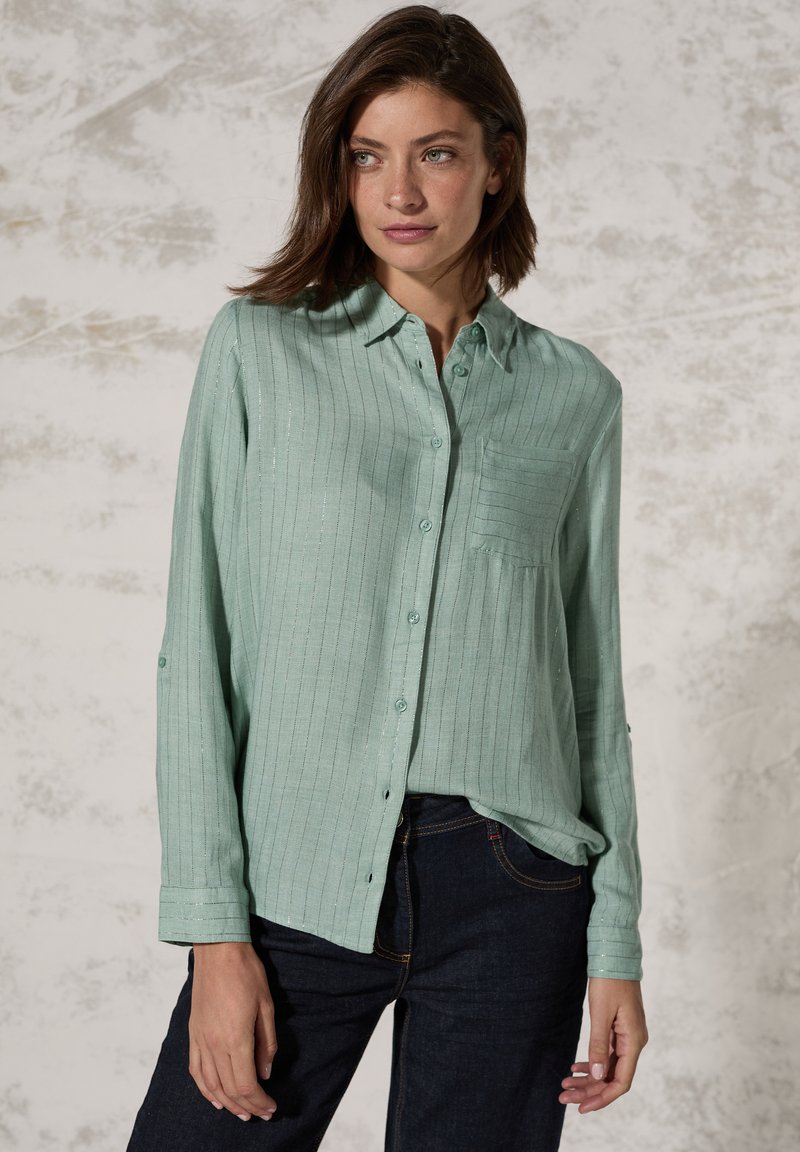 Camicia button-up verde chiaro con righe verticali, una singola tasca sul petto e maniche lunghe arrotolate sui polsi. Realizzata in un tessuto morbido e textured.