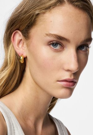 Guldfärgad ringörhänge visad på en modells öra. Örhänget har en slät, rundad design med en polerad yta som reflekterar ljus.