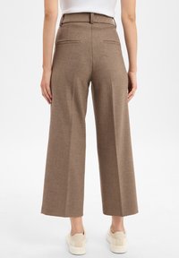 Bruine, wijde broek gemaakt van een gestructureerde stof, met een hoge taille en twee achterzakken. Draagt bij witte sneakers.