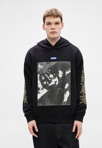 Μαύρο φούτερ με κουκούλα που διαθέτει μεγάλο γραφικό τύπωμα στο μπροστινό μέρος, ετικέτα με λογότυπο και κίτρινα κείμενα στα μανίκια. Πυκνό ύφασμα, χαλαρή εφαρμογή.