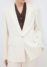 Gräddstripad kavaj med skräddarsydd passform, med en enkel guldknappstängning, slagkrage och två framfickor. Mörkbrun blus under.