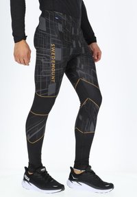 Schwarze Sportleggings mit geometrischem Muster in Grau und Gold, hoher Taillenbund und reflektierenden Akzenten. Getragen mit schwarzen Laufschuhen.