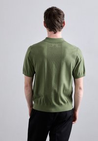 Hombre vestido con polo de punto de manga corta en color verde oliva y pantalones negros, de pie frente a una pared lisa de color gris claro.