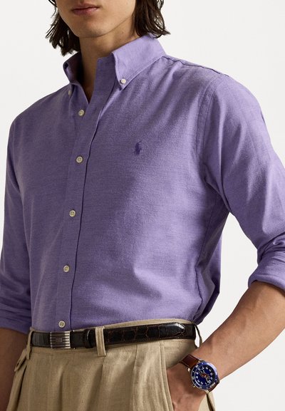 Cămașă button-up de culoare violet deschis, cu guler, având nasturi albi și un mic logo brodat. Purtați cu pantaloni bej și o curea închisă.