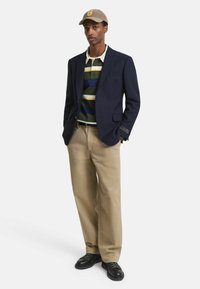 Blazer navy sobre una polo a rayas verde y amarilla, combinado con pantalones beiges y zapatos negros. El conjunto se complementa con una gorra beige.