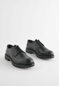 Zapatos de cuero negro con punta redondeada, diseño con cordones y suela de goma gruesa con un patrón de tracción texturizado.