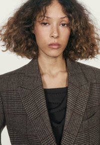 Blazer marrone a quadri con spalle strutturate, rever a punta e una maglia nera testurizzata sotto. Sottile motivo arricchisce il design.