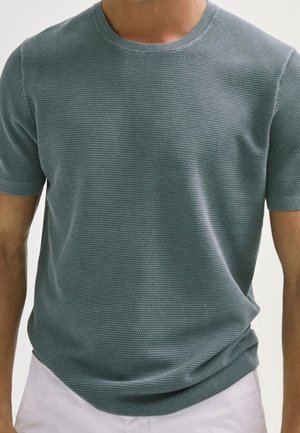 Mann trägt ein kurzärmeliges, strukturiertes, grünes Strickshirt mit rundem Ausschnitt und weißen Hosen, dargestellt von den Schultern bis zur Hüfte.