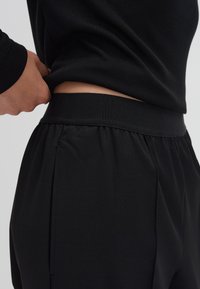 Pantalon de survêtement noir avec une large taille élastique et des poches latérales, présentant une texture lisse et douce ainsi qu'un design plissé.