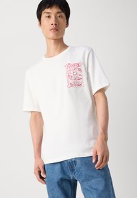 Hvid bomulds T-shirt med korte ærmer og et rødt grafisk design på venstre brystområde, med en stiliseret blomst og tekst.