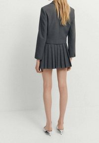 Blondynka stojąca tyłem, ubrana w ciemnoszary krótki żakiet i dopasowaną plisowaną mini spódnicę oraz białe mules na obcasie.