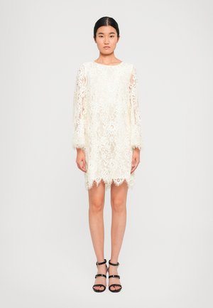 Donna vestita con un abito bianco in pizzo all'altezza del ginocchio, con maniche lunghe e sandali neri con tacco alto e strappi, in piedi contro uno sfondo bianco semplice.