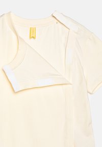 Kremowa koszulka niemowlęca z krótkim rękawem z bocznym zapięciem na zatrzaski i rzepy, zaprojektowana dla łatwego ubierania i zdejmowania.