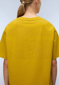 T-shirt jaune surdimensionné en tissu doux. Présente une texture à motifs subtils au dos. Encolure arrondie et manches courtes.