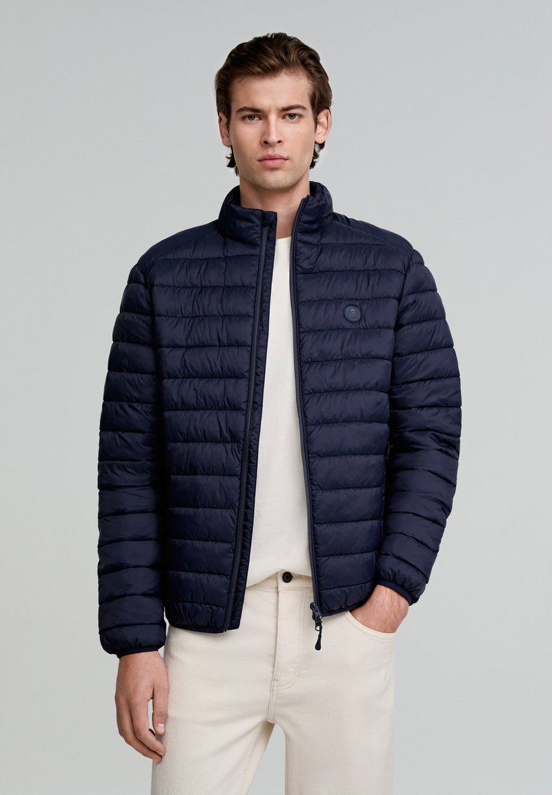 Chaqueta de plumón azul marino con acolchado horizontal, cuello alto, cierre delantero y un pequeño logo. Usada sobre una camisa ligera, combinada con pantalones claros.