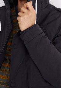 Schwarze Jacke mit Kapuze und weißen Kordeln; verfügt über eine Knopfverschluss, gesteppte Textur und ein sichtbares gestreiftes Hemd darunter.