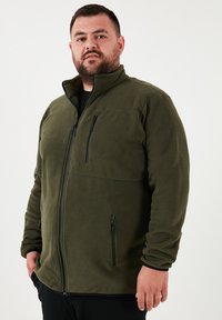 Zelená fleecová bunda s vysokým golierom, plným zipsom a dvoma prednými vreckami na zips. Má hladkú textúru a čierne kovové prvky.