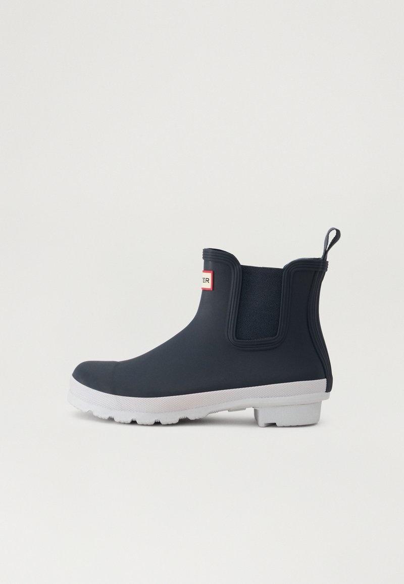 Botte de pluie noire en caoutchouc hauteur cheville avec une semelle blanche et des panneaux élastiques sur les côtés, languette de traction au talon et étiquette de logo à l'avant.