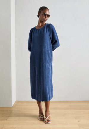Navy blue textured dress with a square neckline, puff sleeves, and two front pockets, complemented by white strappy sandals.