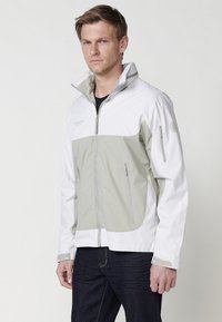 Veste légère en blanc et gris clair, dotée d'une fermeture éclair à l'avant, de poches latérales et d'un col haut. Texture lisse avec des sections contrastantes.