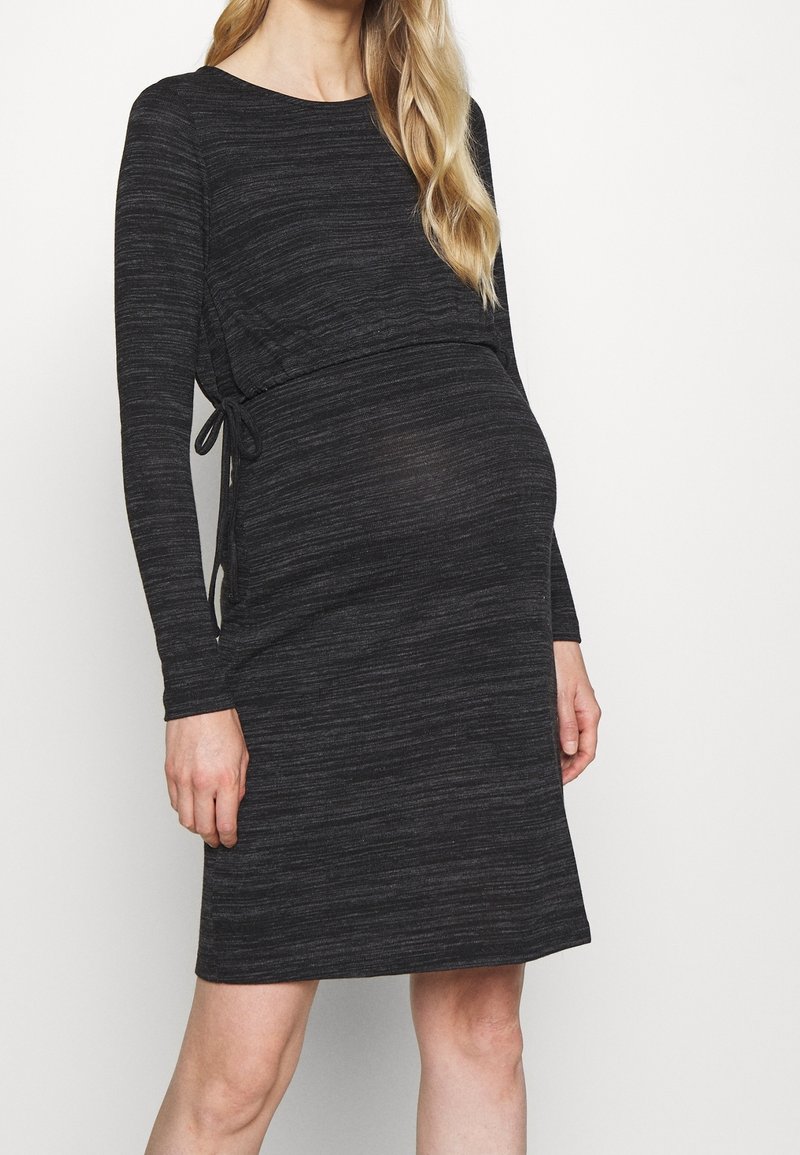 Femme enceinte portant une robe de maternité ajustée, à manches longues, gris foncé, avec un détail de lien sur le côté, sur un fond clair uni.