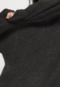 Maglione grigio scuro in maglia realizzato con tessuto morbido, caratterizzato da una texture a coste spesse e un colletto piegato, parzialmente visibile con una mano che afferra la manica.